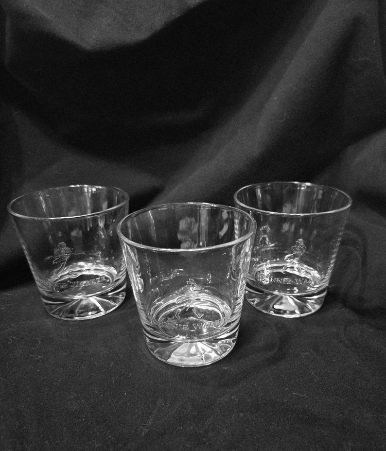 May include: Three clear, lowball whiskey glasses with a rounded base and a slightly flared rim. Each glass has an etched design and the text "JOHNNIE WALKER" on the base. The glasses are arranged on a dark surface.