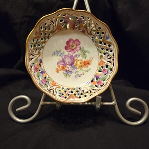 Peut inclure: Petite assiette en porcelaine blanche avec un bord festonné et une bordure dorée. L'assiette présente un motif floral au centre avec des fleurs violettes, roses, jaunes et oranges. Le bord a un design ajouré. L'assiette est présentée sur un présentoir en métal argenté.