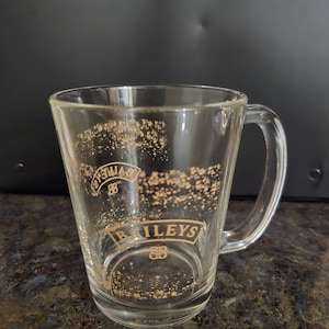 Peut inclure: Mug en verre transparent avec une anse, orné du logo "Baileys" en lettres dorées. Le mug a une forme légèrement évasée et est conçu pour les boissons. Quelques éclaboussures dorées ornent le verre.