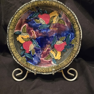 Può includere: Un piatto decorativo in ceramica con un design di melograni dipinto a mano. Il piatto presenta frutti rossi e gialli con foglie blu-verdi su uno sfondo blu e viola. Il bordo ha un bordo decorativo.