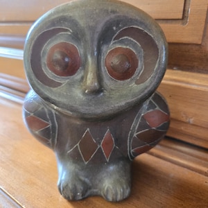 May include: A ceramic owl ornament with a grey body and large, round eyes. The eyes are brown with concentric circles. The wings feature a geometric pattern of red and brown diamonds. The owl is a decorative item.