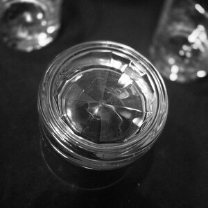 May include: A black and white image of a clear, faceted glass tumbler. The glass is in focus, with two other out-of-focus glasses in the background. The glass sits on a dark surface, reflecting light.