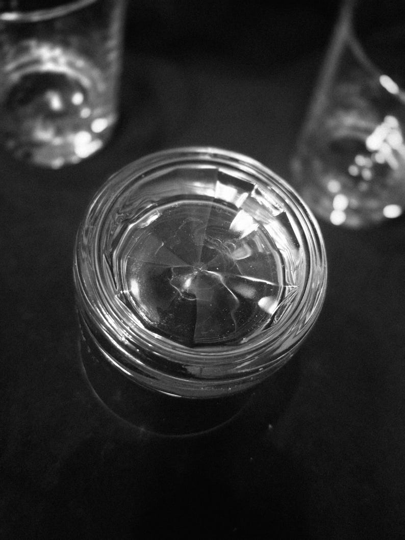 May include: A black and white image of a clear, faceted glass tumbler. The glass is in focus, with two other out-of-focus glasses in the background. The glass sits on a dark surface, reflecting light.