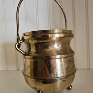 May include: A small, gold-toned metal pot with a handle and three legs. The pot has a rounded body and a smooth, shiny finish.