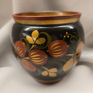 May include: A hand-painted, round wooden bowl with a black base and a decorative floral design. The bowl features red, yellow, and green painted flowers and leaves. The rim is trimmed with red and gold paint.