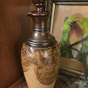 May include: A tall, handcrafted ceramic bottle with a stopper. The bottle features a brown and tan colour scheme with a textured, speckled design. The stopper is a rounded shape. The bottle is displayed near a framed picture.