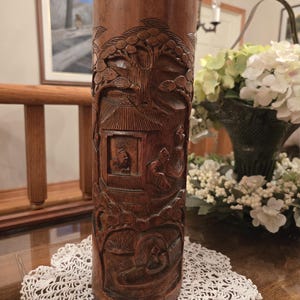 May include: A cylindrical, dark brown wooden vase with detailed carvings of a landscape scene. The carvings feature trees, buildings, and figures. The vase is displayed on a white, lace doily.