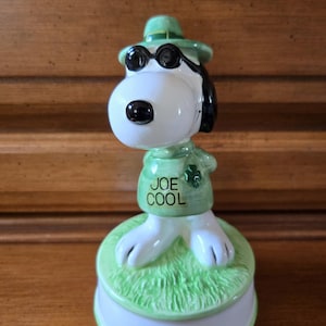 May include: A ceramic Snoopy figurine in a green hat and sunglasses, with the words "JOE COOL" on his shirt. The figurine is on a green base with a white border, and the overall design is a playful and collectible item.