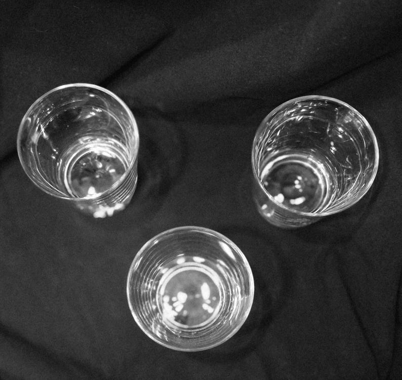May include: Three clear, empty drinking glasses arranged on a dark surface. The glasses are cylindrical with a slightly textured exterior. The image is a high-angle shot, showcasing the glasses' interiors.