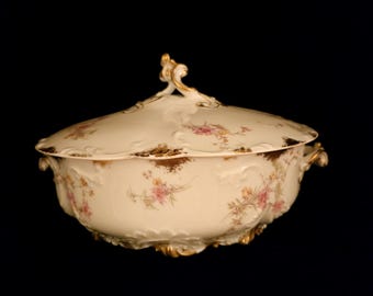 May include: A white porcelain serving bowl with a lid, featuring a floral pattern in shades of pink and gold. The bowl has a scalloped edge and ornate handles.