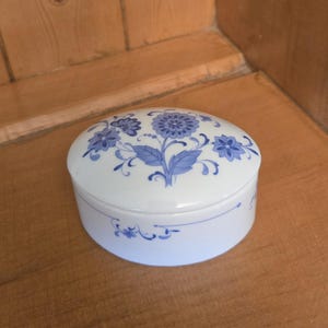 Peut inclure: Boîte à bijoux ovale en porcelaine blanche, ornée d'un motif floral bleu sur le couvercle et les côtés. Le motif représente des fleurs et des feuilles stylisées. Posée sur une surface en bois.
