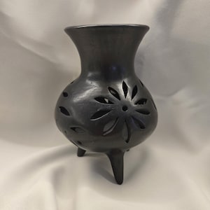 May include: A dark grey, handcrafted ceramic vase with a bulbous body and three legs. The vase features decorative cut-out floral patterns and a flared top. The vase is approximately 15 cm tall.