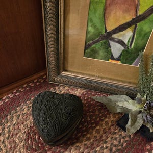 Puede incluir: Una caja negra en forma de corazón con intrincados patrones florales y geométricos tallados. La caja está sobre una alfombra trenzada con tonos rojos y marrones, cerca de una acuarela enmarcada de un pájaro.