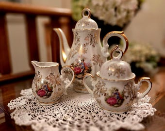 May include: A white porcelain tea set with gold trim, featuring a teapot, creamer, and sugar bowl. Each piece is decorated with floral and figural designs in shades of pink, red, and blue. The set rests on a white lace doily.