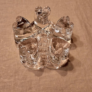 Könnte beinhalten: Ein durchsichtiger Kerzenhalter aus Glas mit fünf herzförmigen Kerzenschalen. Der Kerzenhalter besteht aus klarem Glas und steht auf einer strukturierten Oberfläche. Das Design ist symmetrisch und dekorativ.