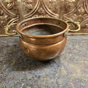 May include: A small copper pot with two handles. The pot is round and has a slightly flared lip. The pot is made of copper and has a shiny finish.