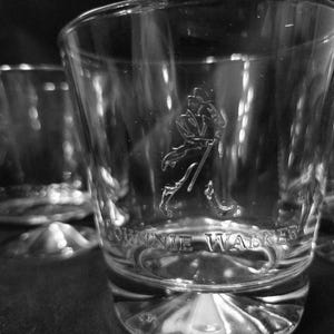 May include: Close-up of three clear, etched whiskey glasses. The glasses feature the Johnnie Walker logo and text. The glasses have a thick base and a slightly flared rim. The image is in black and white.