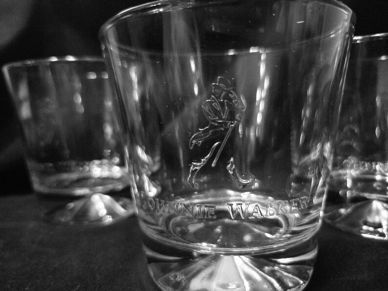 May include: Close-up of three clear, etched whiskey glasses. The glasses feature the Johnnie Walker logo and text. The glasses have a thick base and a slightly flared rim. The image is in black and white.
