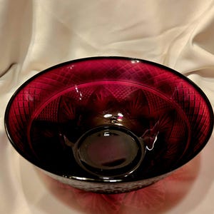 May include: A deep red glass bowl with a textured, diamond-patterned exterior and interior. The bowl has a wide, oval shape and a smaller, circular base inside. The glass is transparent, allowing light to pass through and highlight the intricate design.