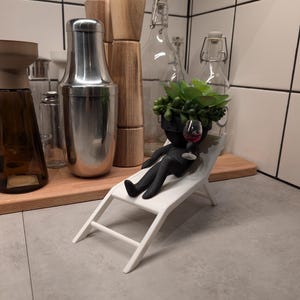 May include: A black figurine of a person relaxing on a white lounge chair, holding a glass of red wine. The figurine has a succulent plant growing out of its head.