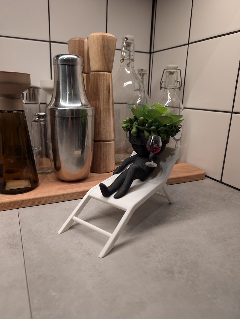 May include: A black figurine of a person relaxing on a white lounge chair, holding a glass of red wine. The figurine has a succulent plant growing out of its head.