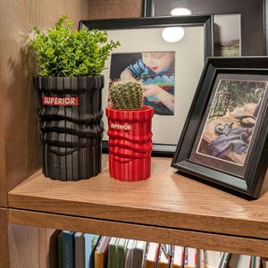 May include: Two black and red plant pots with the word "SUPERIOR" printed on them. The pots are on a wooden shelf with framed pictures and books behind them.