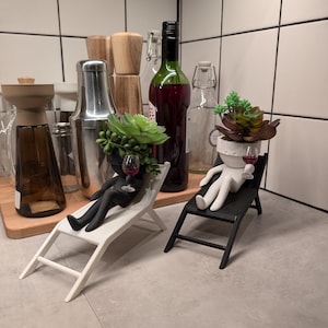 May include: Two black and white figurines of people sitting in lounge chairs. Each figurine has a succulent plant growing out of their head and is holding a small glass of red wine. The figurines are on a gray tiled surface.