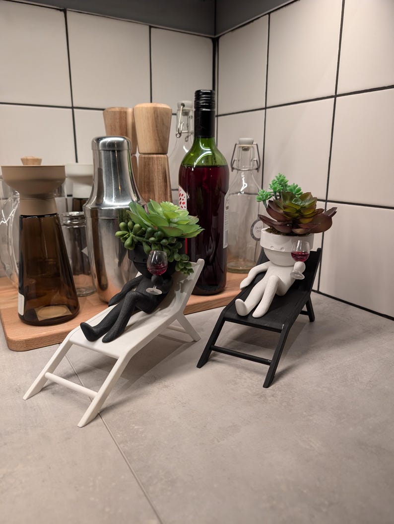 May include: Two black and white figurines of people sitting in lounge chairs. Each figurine has a succulent plant growing out of their head and is holding a small glass of red wine. The figurines are on a gray tiled surface.