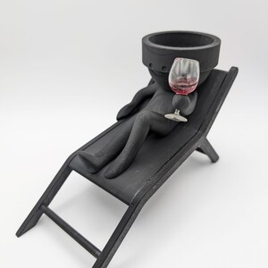May include: Black ceramic figurine of a person sitting on a black wooden lounge chair, holding a glass of red wine.