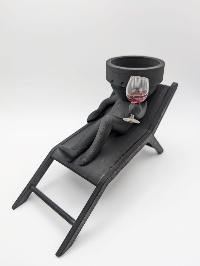 May include: Black ceramic figurine of a person sitting on a black wooden lounge chair, holding a glass of red wine.