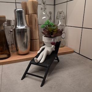 May include: A white ceramic figurine of a person sitting in a black lounge chair, holding a small glass of red wine. The figurine has a potted succulent plant for a head.