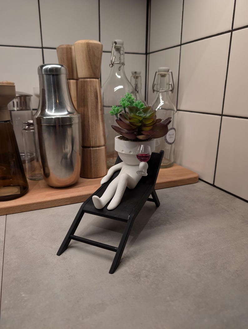 May include: A white ceramic figurine of a person sitting in a black lounge chair, holding a small glass of red wine. The figurine has a potted succulent plant for a head.