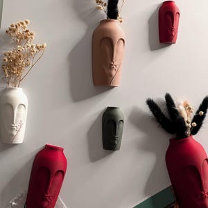 May include: Five ceramic vases in different colours, each shaped like a face with a single stem of dried flowers in the top. The vases are mounted on a white wall.