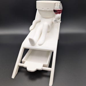 May include: White figurine of a person relaxing on a lounge chair with a glass of red wine. The figurine has a small drawer underneath the chair.