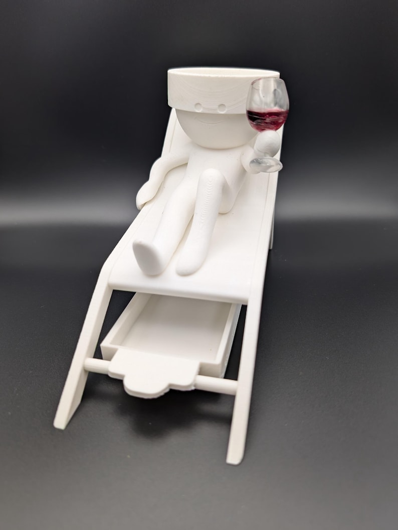 May include: White figurine of a person relaxing on a lounge chair with a glass of red wine. The figurine has a small drawer underneath the chair.