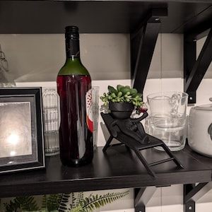 May include: A black shelf with a bottle of red wine, a small potted succulent plant, and a black figurine of a person sitting on a lounge chair.