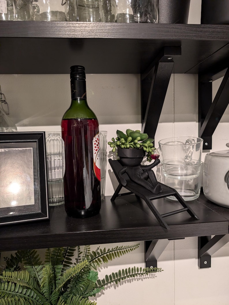 May include: A black shelf with a bottle of red wine, a small potted succulent plant, and a black figurine of a person sitting on a lounge chair.