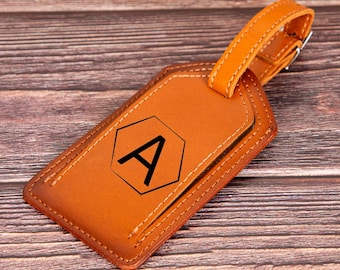 May include: A brown leather luggage tag with a stitched border and a black letter "A" inside a hexagon. The tag has a brown leather strap with a metal buckle.