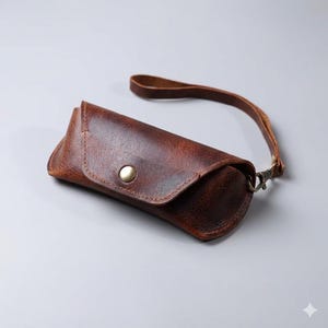 May include: A brown leather sunglasses case with a wrist strap. The case has a rounded shape with a snap closure. The leather has a distressed appearance, and the strap is attached with a metal clasp.