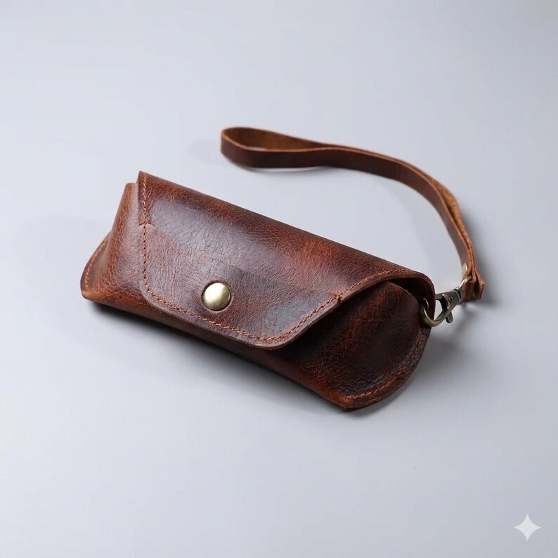 May include: A brown leather sunglasses case with a wrist strap. The case has a rounded shape with a snap closure. The leather has a distressed appearance, and the strap is attached with a metal clasp.