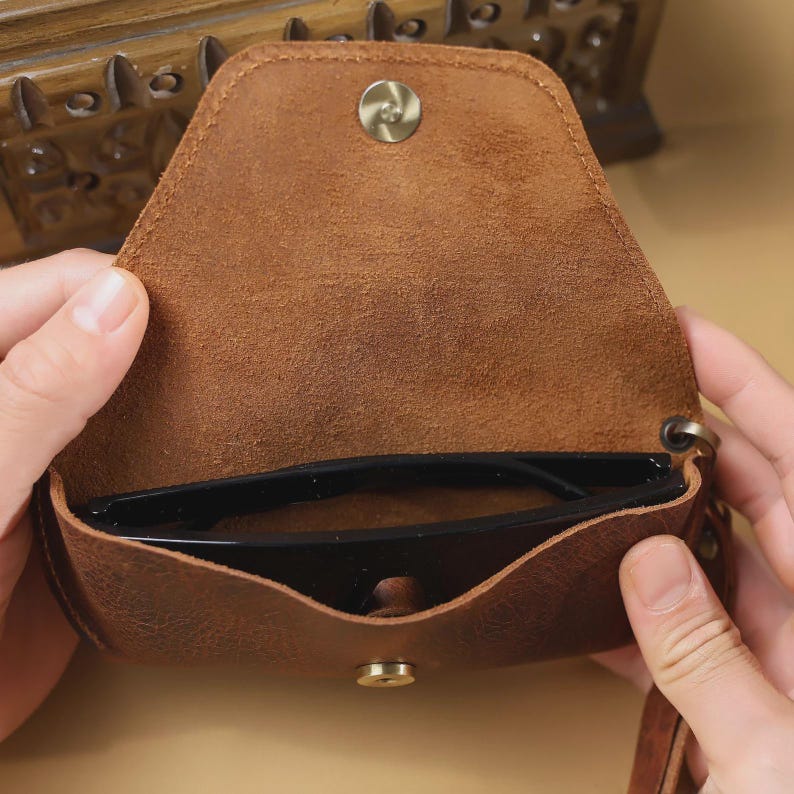 May include: A brown leather glasses case, open to reveal a pair of black sunglasses. The case has a snap closure and a small strap. The leather has a distressed appearance.