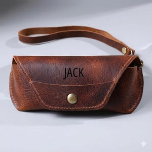 May include: A brown leather glasses case with the name "JACK" embossed on the front. The case has a snap closure and a wrist strap. The leather has a distressed look.