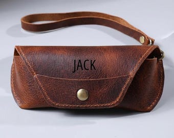 May include: A brown leather glasses case with the name "JACK" embossed on the front. The case has a snap closure and a wrist strap. The leather has a distressed look.