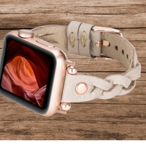 May include: A rose gold Apple Watch with a white braided leather band. The watch face displays a photo of a canyon.