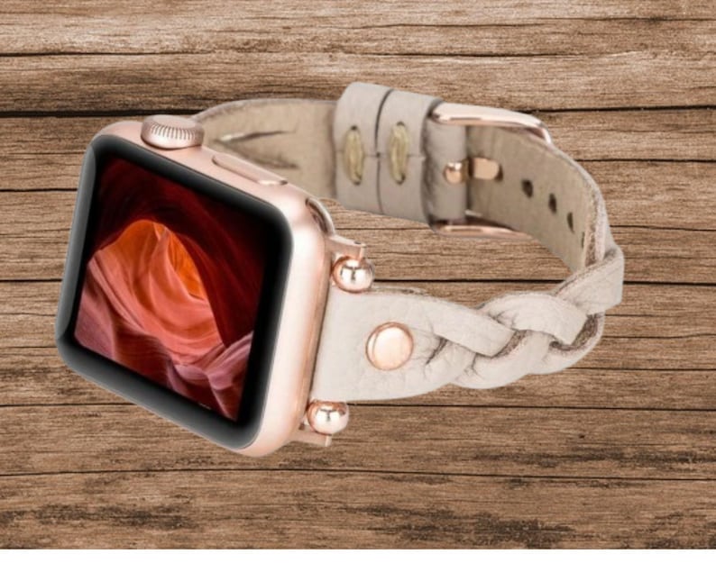May include: A rose gold Apple Watch with a white braided leather band. The watch face displays a photo of a canyon.