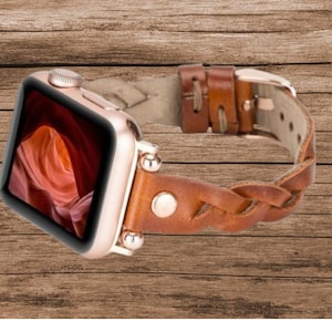 May include: A brown leather braided Apple Watch band with a rose gold buckle. The band is woven with a braided pattern and has a smooth leather finish.