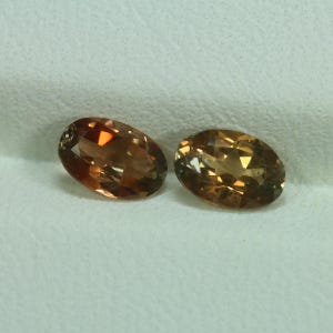 May include: Two oval-shaped gemstones with a warm, golden-brown hue. The gems have a polished surface that reflects light, creating a sparkling effect. The stones are set against a soft, white background.
