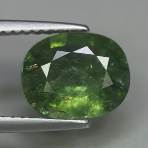 May include: A faceted, oval-shaped green gemstone held by a pair of tweezers. The gem has a vibrant, translucent appearance with internal inclusions. The cut features multiple facets that reflect light, creating a sparkling effect. The tweezers are silver.
