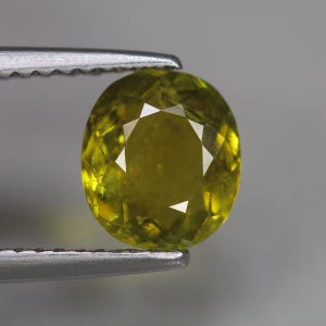 May include: An oval-cut, faceted gemstone held by a pair of silver tweezers. The gem is a vibrant yellow-green color, with multiple facets creating a sparkling effect. The background is a soft gray.