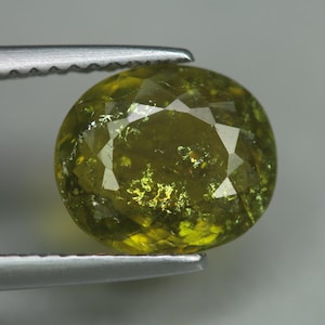 May include: A faceted, oval-shaped gemstone held by tweezers. The gem is a vibrant olive green color with internal inclusions and a brilliant, reflective surface. The facets catch the light, creating a sparkling effect.